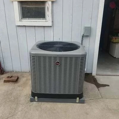 Gray central air conditioning condenser unit installed on concrete pad against white siding
